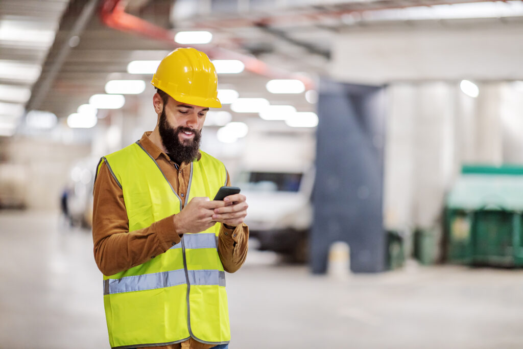 Ein Bauarbeiter mit gelbem Sicherheitshelm und reflektierender Weste überprüft sein Smartphone auf der Baustelle und lächelt.